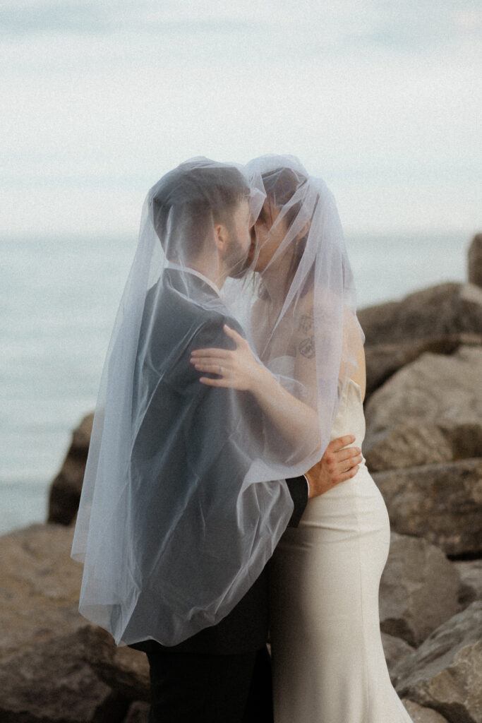 Bride and groom sharing an intimate kiss under bride veil, overlooking Lake Ontario on their wedding day